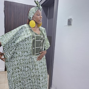 May include: A floor-length olive green kaftan featuring a white geometric pattern throughout, a solid olive green V-neck panel with white embroidered line details, a matching olive green headwrap with a white pattern, and large yellow circular earrings.