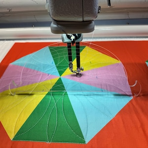 May include: A close-up of a sewing machine quilting a colorful fabric with a circular design. The fabric is made up of six triangles in different colors, including pink, yellow, green, and blue. The sewing machine is using a clear plastic template to guide the stitching.