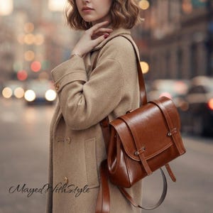 Peut inclure: Un sac à dos en cuir marron avec un rabat, deux sangles verticales à boucles métalliques et des bretelles réglables. Le sac est porté sur un manteau en laine beige avec des boutons visibles. Le texte Mayed with Style est présent.