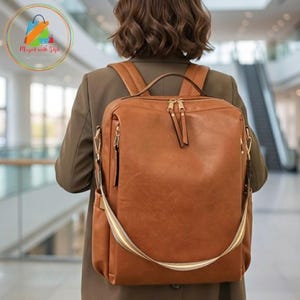 May include: A tan rectangular backpack with a smooth finish, featuring a top handle, two shoulder straps, and gold-toned metal zippers. The bag has a large main compartment with a front pocket and side pockets, accented by a light-colored strap detail.