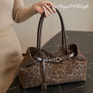 May include: A brown leopard print tote bag featuring dark brown leather-like handles and a matching thin belt detail with a silver-toned buckle across the front. The bag has a soft, textured surface and includes the text MayedWithStyle.