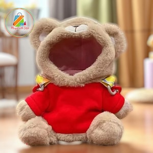 May include: A plush teddy bear wearing a red hooded sweatshirt with a yellow and patterned collar. The bear has a light brown fur and a white snout. The image has a soft focus.
