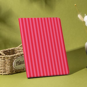 Peut inclure: Une housse de tablette à rayures roses et rouges est posée sur une surface verte. Un panier en osier avec une étiquette de plante est à gauche. Un vase avec des fleurs séchées est à droite. Le mot "Shadeify" est en bas.