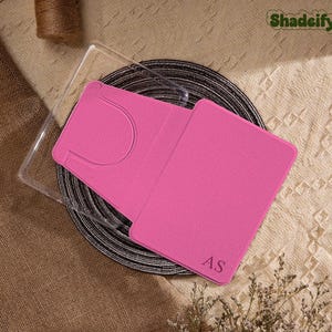 Könnte beinhalten: Ein leuchtend rosa Kartenhalter mit den Initialen "AS" wird offen gezeigt und zeigt eine Tasche. Der Kartenhalter befindet sich in einer transparenten Kunststoffhülle und liegt auf einer schwarz-grauen gewebten Matte. Das Wort "Shadeify" ist oben rechts sichtbar.