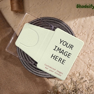 May include: A light green Kindle case with the text "YOUR IMAGE HERE" and "Customized images for your kindle". The case is on a dark brown circular object, with a clear plastic case and a brown twine roll in the background.