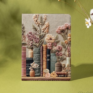 Pode incluir: Uma capa decorativa para tablet com um design bordado de livros, vasos e flores em tons de rosa, bege e verde. A capa tem uma aparência texturizada e um detalhe de prateleira de madeira. A marca "Shadeify" é visível no canto inferior direito.