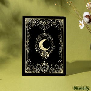 Pode incluir: Diário preto com um design floral e de lua crescente dourada. A capa apresenta uma ilustração detalhada de uma lua crescente, uma rosa e elementos botânicos ornamentados. O diário está sobre um fundo verde.