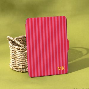 Puede incluir: Una funda para tableta a rayas rosas y rojas con las iniciales "MK" en dorado. Una cesta de mimbre se encuentra a la izquierda de la funda. El fondo es de color verde liso. La marca "Shadeify" está en la esquina inferior derecha.