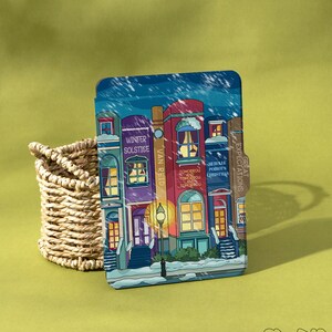 以下が含まれることがあります： 建物、雪、そして「Winter Solstice」と「Great Expectations」のテキストが描かれた冬のシーンの装飾カード。 カードは、緑色の表面にある編まれたバスケットの隣に配置されています。