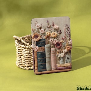 以下が含まれることがあります： 花と本のデザインが施された刺繍タブレットケース。ケースはベージュの背景にカラフルな花と本の山があります。織りのバスケットがケースの左側にあります。「Shadeify」のブランド名は右下にあります。