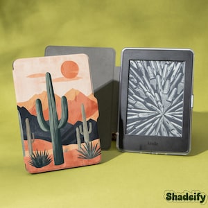 Peut inclure: Une liseuse Kindle avec une couverture représentant un paysage désertique avec des cactus, des montagnes et un soleil couchant. La couverture est beige, orange et verte. La liseuse est noire avec un étui de protection transparent. La marque "Shadeify" est en bas à droite.
