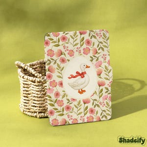 Könnte beinhalten: Eine Tablet-Hülle mit floralem Muster und einer Cartoon-Ente mit roter Schleife. Die Hülle ist mit rosa Blumen und grünen Blättern verziert. Links befindet sich ein Weidenkorb. Der Markenname "Shadeify" ist unten rechts sichtbar.