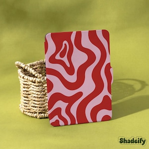 Könnte beinhalten: Eine Tablet-Hülle mit rotem und rosa Muster und einem wellenförmigen Design. Die Hülle steht neben einem Weidenkorb auf einer gelb-grünen Oberfläche. Der Markenname "Shadeify" ist in grüner Schrift gedruckt.
