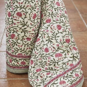 May include: A quilted textile featuring an off-white base with a repeating pattern of dark green vines and pink floral blossoms. The fabric includes a horizontal border with a pink floral band and a thin dark green striped edge at the bottom.