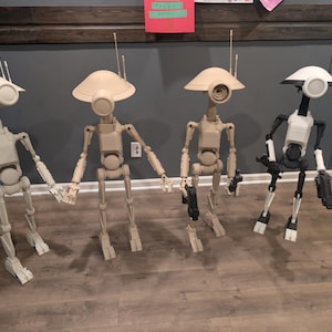 May include: Four droid figures with antenna, in various shades of beige and black. The droids have a rounded head and a cylindrical body. Two droids are holding hands. The droids are standing on a wooden floor.