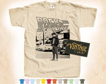 Puede incluir: Una camiseta beige de aspecto vintage con un gráfico de Marty McFly de la película "Regreso al futuro". El gráfico muestra a Marty de pie frente a un DeLorean, una máquina del tiempo, con el texto "Regreso al futuro" sobre él. La camiseta también tiene una etiqueta vintage "Vintage T-Shirt".