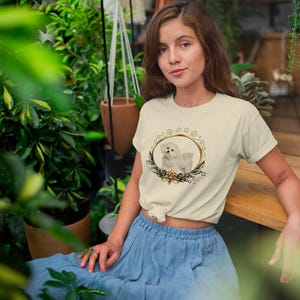 May include: Cream-colored t-shirt featuring a circular design with a white dog illustration inside a gold frame, surrounded by floral accents. The person is wearing a blue skirt and is sitting on a wooden bench, surrounded by plants.
