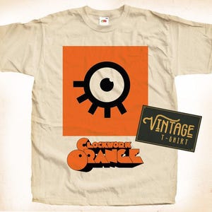 Może przedstawiać: Beżowa koszulka z nadrukiem w stylu vintage czarnego oka z białą źrenicą i tekstem "Clockwork Orange" w pomarańczowych literach. Koszulka ma również vintage'ową etykietę z napisem "Vintage T-Shirt".
