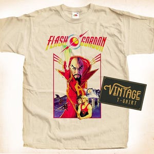 Può includere: Una maglietta vintage beige con una stampa grafica di Flash Gordon, un personaggio del film degli anni '80. La grafica presenta Flash Gordon con un mantello rosso e una donna in un abito verde e bianco. Il testo "Flash Gordon" è stampato in nero e rosso.