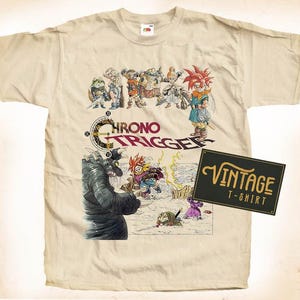 May include: A beige t-shirt with a vintage look featuring a graphic of characters from the video game Chrono Trigger. The graphic includes the game's title and a group of characters in a battle scene. The t-shirt has a vintage t-shirt label attached to the bottom right corner.
