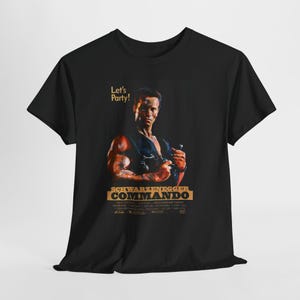 May include: Black t-shirt featuring a movie poster design. The design includes an image of Arnold Schwarzenegger and the text "SCHWARZENEGGER COMMANDO" in gold lettering. The phrase "Let's Party!" is also present.