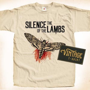 Op de afbeelding: Een beige t-shirt met een vintage t-shirt label. Het shirt heeft een afbeelding van een nachtvlinder met een schedelkop en bloed dat van zijn vleugels druppelt. De tekst "Silence of the Lambs" is boven de nachtvlinder gedrukt.