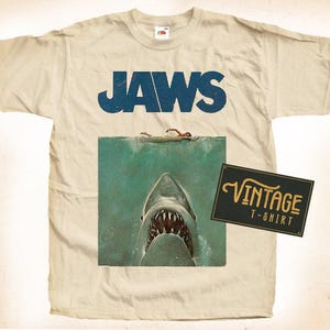 Pode incluir: Uma camiseta vintage bege com um gráfico azul "JAWS" e uma ilustração vintage de um tubarão. A camiseta também tem uma etiqueta "Vintage T-Shirt".