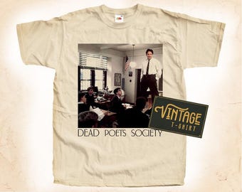 May include: A beige vintage t-shirt with a print of a classroom scene and the text "Dead Poets Society". The print features a group of students and a teacher standing in front of a chalkboard. The t-shirt also has a vintage label that says "Vintage T-Shirt".