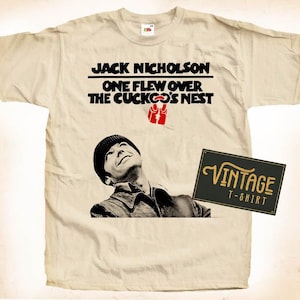 Puede incluir: Una camiseta beige de aspecto vintage con una imagen en blanco y negro de Jack Nicholson de la película "Atrapado sin salida". El texto "Jack Nicholson One Flew Over The Cuckoo's Nest" está impreso en negro en la camiseta. La camiseta también tiene una etiqueta vintage "T-shirt".