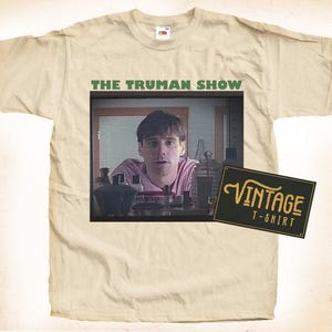 Op de afbeelding: Een beige t-shirt met een vintage look met een screenshot van Jim Carrey uit de film "The Truman Show" en de tekst "The Truman Show" boven de afbeelding. De tekst "Vintage T-Shirt" staat op een zwarte rechthoek in de rechterbenedenhoek van de afbeelding.