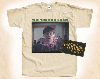 Könnte beinhalten: Ein beigefarbenes T-Shirt mit Vintage-Look, das einen Screenshot von Jim Carrey aus dem Film "The Truman Show" und den Text "The Truman Show" über dem Bild zeigt. Der Text "Vintage T-Shirt" befindet sich auf einem schwarzen Rechteck in der unteren rechten Ecke des Bildes.