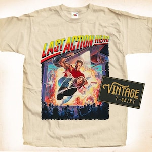 Pode incluir: Uma camiseta bege com um visual vintage com um gráfico de Arnold Schwarzenegger como Jack Slater do filme "O Último Herói da Ação". O gráfico mostra Slater no ar, segurando uma arma, com uma cidade ao fundo. O texto "Last Action Hero" está impresso em letras negritas acima do gráfico.