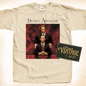 May include: A vintage white t-shirt with a print of the movie "Devil's Advocate" featuring Al Pacino and Keanu Reeves. The print shows the two actors in suits, with Pacino standing behind Reeves and placing a hand on his shoulder. The t-shirt has a vintage label that says "Vintage T-Shirt".