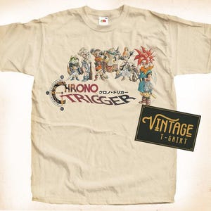 May include: A vintage beige t-shirt with a graphic print of characters from the video game Chrono Trigger. The graphic includes the game's logo and the Japanese text "Chrono Trigger". The t-shirt has a vintage look and feel.