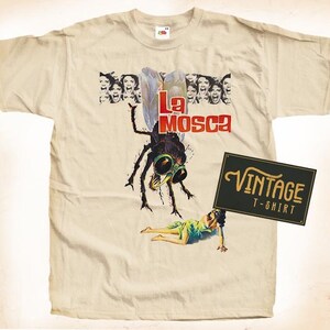 May include: A vintage beige t-shirt featuring a graphic of a large fly with the text "La Mosca" printed on it. The fly is depicted in a menacing way with sharp claws and a large eye. The t-shirt also features a vintage label that says "Vintage T-Shirt".