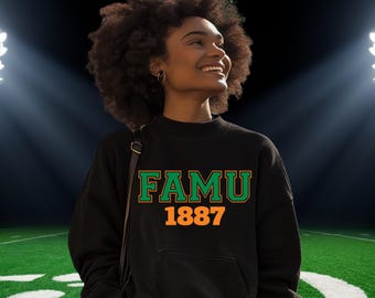 May include: Black sweatshirt with the text "FAMU 1887" in green and orange. The sweatshirt has a front pocket and a crew neck. The background shows a football field with stadium lights.
