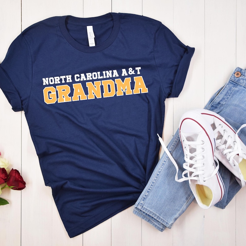 May include: Navy blue t-shirt with the text "NORTH CAROLINA A&T GRANDMA" in white and gold lettering. The shirt is paired with blue jeans and white sneakers with red accents, arranged on a white wooden surface.