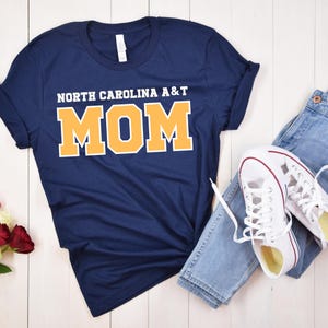 May include: Navy blue t-shirt with the text "NORTH CAROLINA A&T MOM" in white and gold lettering. The shirt is paired with blue jeans and white sneakers with red accents, creating a casual, everyday look.