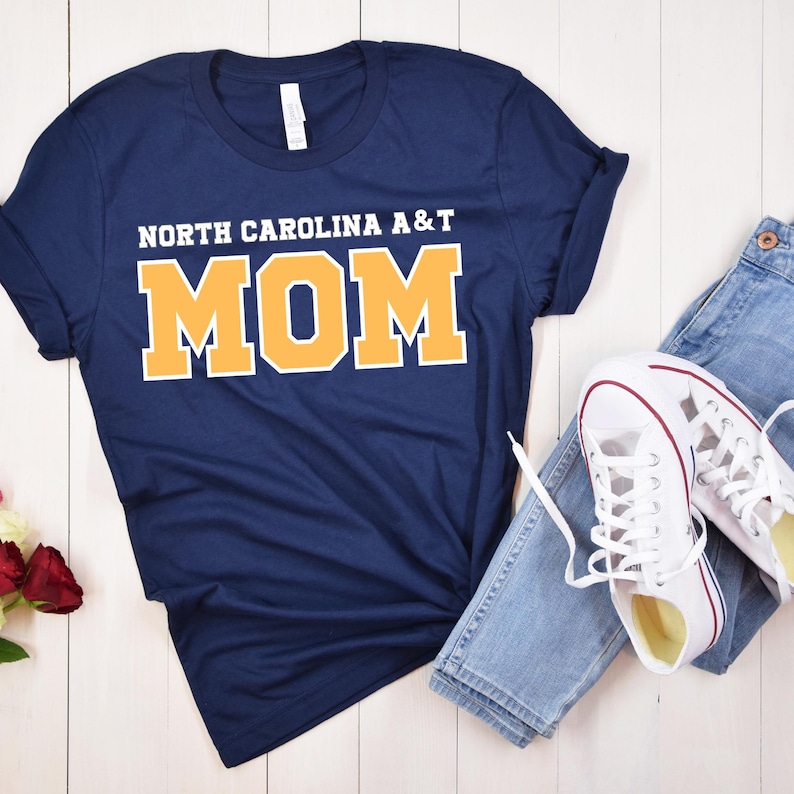 May include: Navy blue t-shirt with the text "NORTH CAROLINA A&T MOM" in white and gold lettering. The shirt is paired with blue jeans and white sneakers with red accents, creating a casual, everyday look.