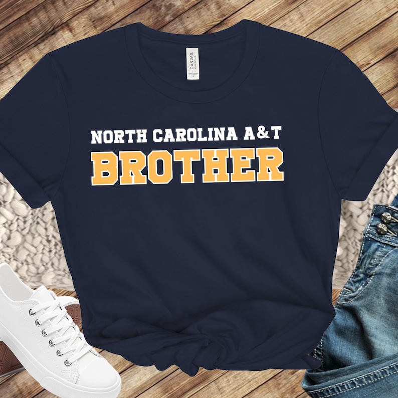May include: Navy blue t-shirt with the text "NORTH CAROLINA A&T BROTHER" in white and orange block letters. The shirt is made of a soft material and has short sleeves.