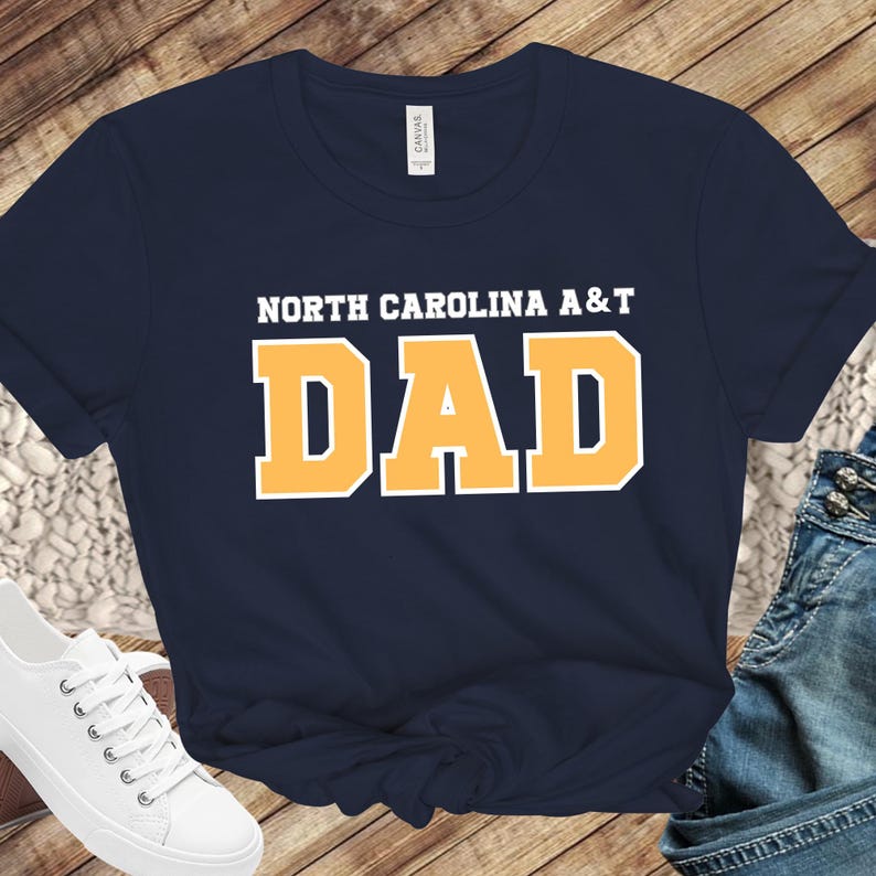 May include: Navy blue t-shirt with the text "NORTH CAROLINA A&T DAD" in white and gold lettering. The shirt is a classic crew neck style and is paired with jeans and white sneakers.