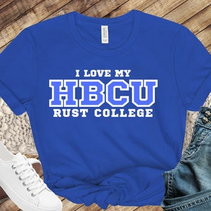 May include: Royal blue t-shirt with the text "I LOVE MY HBCU RUST COLLEGE" in white and blue lettering. The shirt is displayed on a wooden surface with a pair of white sneakers and denim jeans.