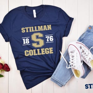 May include: Navy blue t-shirt with "STILLMAN COLLEGE" printed in gold and white. The shirt features the year "1876" and the letter "S" in a collegiate design. The shirt is paired with blue jeans and white sneakers.
