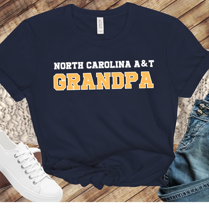 May include: Navy blue t-shirt with the text "NORTH CAROLINA A&T GRANDPA" in white and orange block letters. The shirt is made of a soft material and has a classic crew neck design.