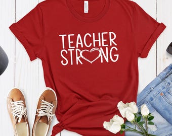 May include: A red short-sleeved t-shirt featuring the white text "TEACHER STRONG" with a heart symbol replacing the letter "O" in the word "STRONG". The shirt has a crew neckline and is displayed alongside brown sneakers and white roses.