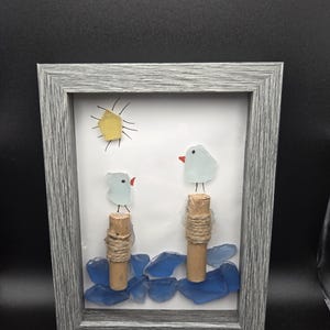 Puede incluir: Obra de arte enmarcada que presenta una escena playera caprichosa. Dos pájaros de cristal azul claro con picos rojos se posan sobre postes de corcho envueltos en hilo, en medio de 'olas' de cristal azul. Un sol amarillo con rayos negros brilla arriba. El marco gris añade un toque rústico.