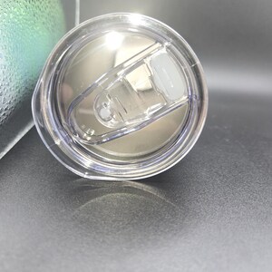 K&ouml;nnte beinhalten: Transparenter Kunststoff-Becherdeckel mit Schiebeverschluss. Der Deckel ist kreisf&ouml;rmig und hat eine silberne Innenseite. Der Schiebeverschluss ist wei&szlig; und in geschlossener Position. Der Deckel liegt auf einer dunkelgrauen Oberfl&auml;che.