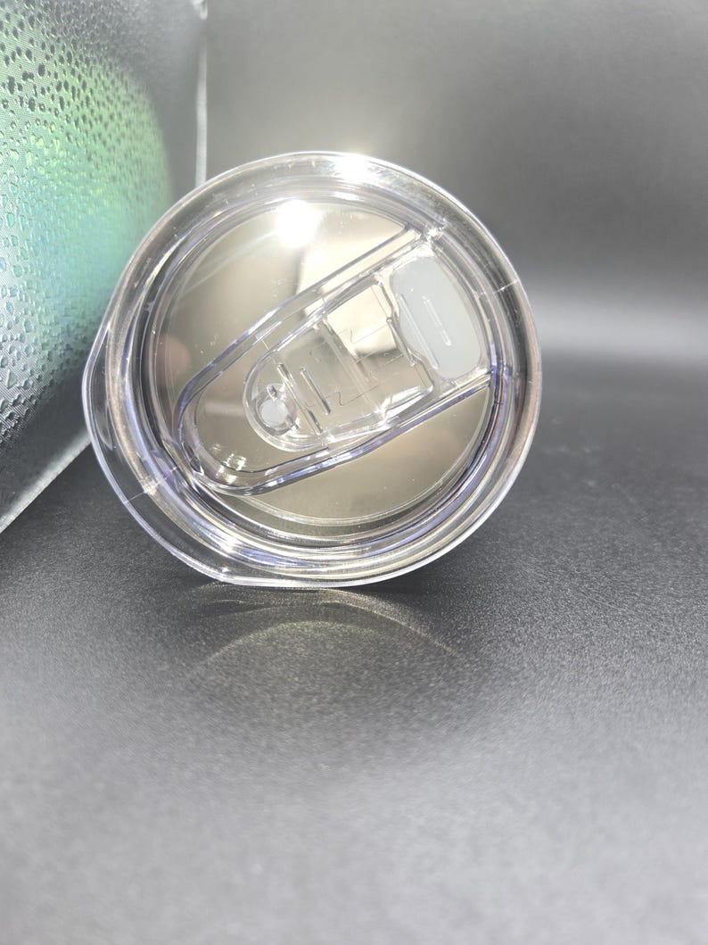 K&ouml;nnte beinhalten: Transparenter Kunststoff-Becherdeckel mit Schiebeverschluss. Der Deckel ist kreisf&ouml;rmig und hat eine silberne Innenseite. Der Schiebeverschluss ist wei&szlig; und in geschlossener Position. Der Deckel liegt auf einer dunkelgrauen Oberfl&auml;che.