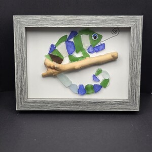 May include: A framed artwork featuring a chameleon made from colorful sea glass pieces. The chameleon is green, blue, and white and is perched on a branch. The artwork is set against a white background.