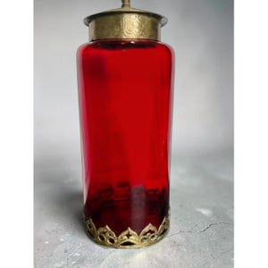 May include: A tall cylindrical glass vessel in a deep red hue, featuring a metallic gold-toned lid with an etched pattern and a decorative gold-toned base with a scalloped, openwork design that encircles the bottom of the glass container.
